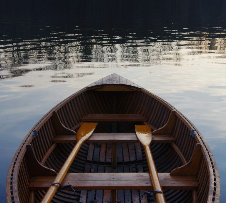 Paarberatung, Einzelberatung, Eheberatung Frankfurt Holzboot mit Paddeln auf ruhigem Wasser, umgeben von Bäumen und reflektierendem Himmel.